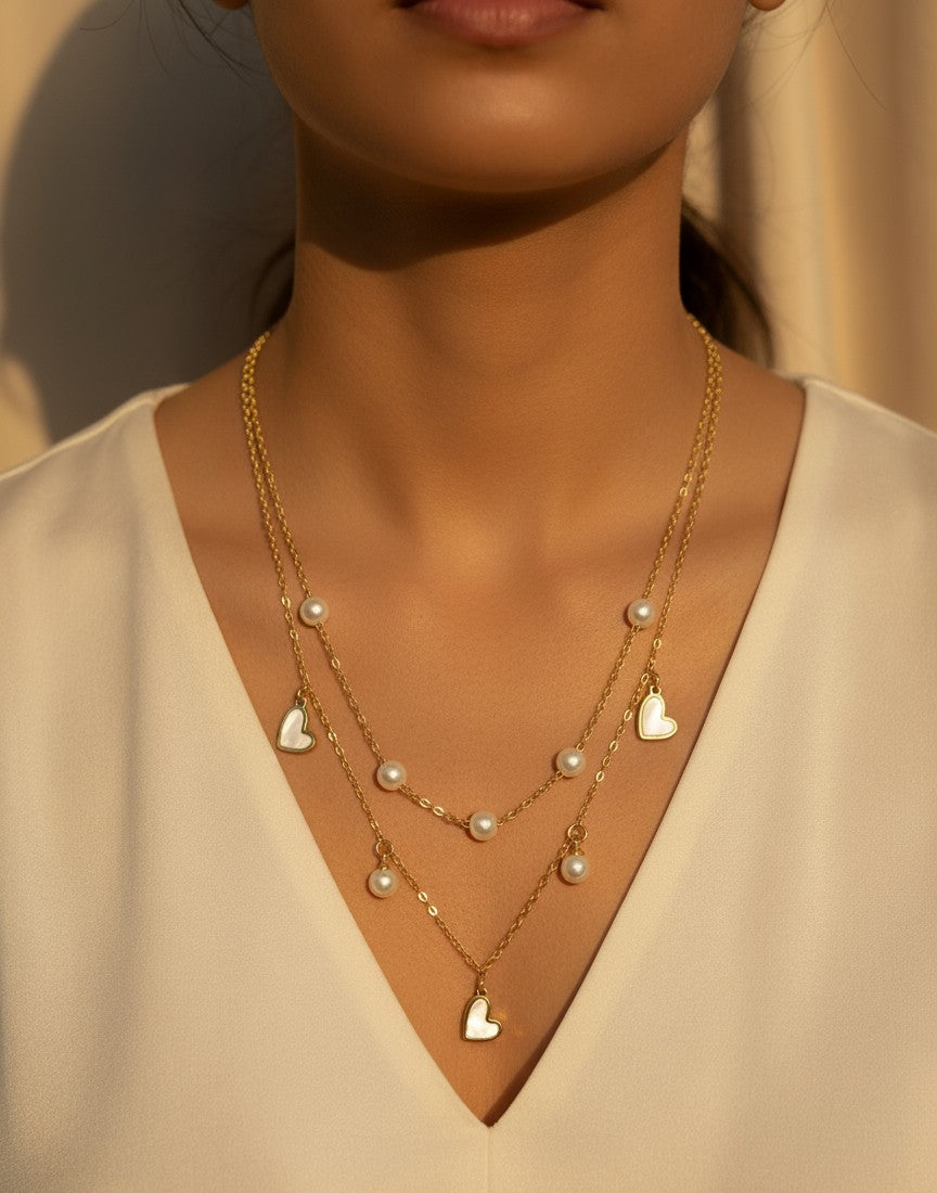 Woman wearing a gold necklace with heart and pearl-shaped pendants on a neutral background